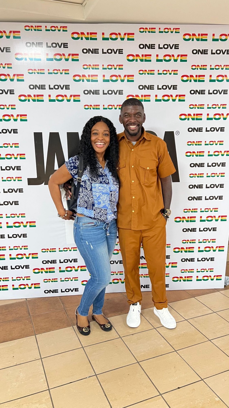 The couple smiling in front of a One Love backdrop